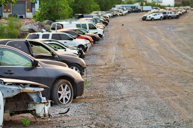 A row of parked cars on a gravel lot with trees in the background. Some cars appear to be damaged or missing parts, and the area has an industrial feel with a warehouse visible at the edge.