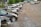 A row of parked cars on a gravel lot with trees in the background. Some cars appear to be damaged or missing parts, and the area has an industrial feel with a warehouse visible at the edge.