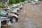 A row of parked cars on a gravel lot with trees in the background. Some cars appear to be damaged or missing parts, and the area has an industrial feel with a warehouse visible at the edge.
