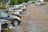 A row of parked cars on a gravel lot with trees in the background. Some cars appear to be damaged or missing parts, and the area has an industrial feel with a warehouse visible at the edge.
