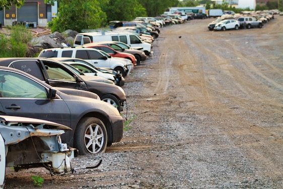 A row of parked cars on a gravel lot with trees in the background. Some cars appear to be damaged or missing parts, and the area has an industrial feel with a warehouse visible at the edge.