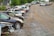 A row of parked cars on a gravel lot with trees in the background. Some cars appear to be damaged or missing parts, and the area has an industrial feel with a warehouse visible at the edge.