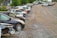 A row of parked cars on a gravel lot with trees in the background. Some cars appear to be damaged or missing parts, and the area has an industrial feel with a warehouse visible at the edge.