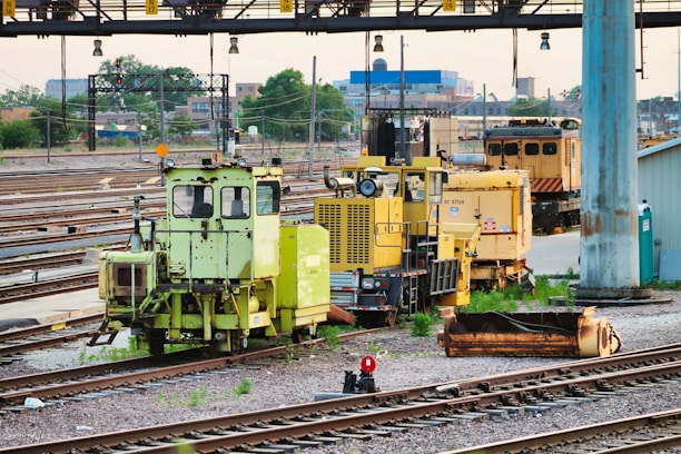 Numerous maintenance vehicles and equipment are stationed on railway tracks. The area appears to be an industrial or depot setting with multiple rail lines converging. Various colors of machinery, predominantly yellow and green, are visible, and some construction or maintenance materials are scattered nearby.