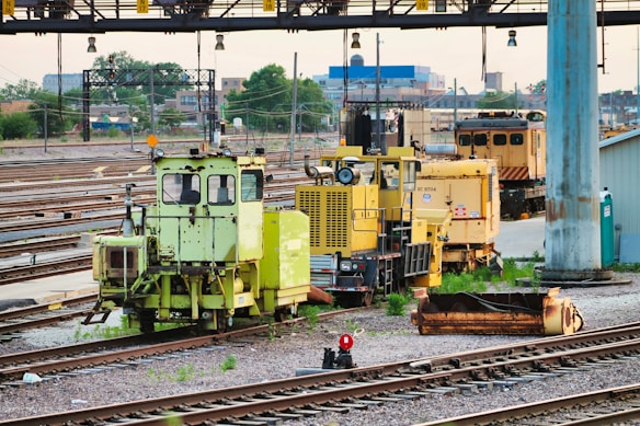 Numerous maintenance vehicles and equipment are stationed on railway tracks. The area appears to be an industrial or depot setting with multiple rail lines converging. Various colors of machinery, predominantly yellow and green, are visible, and some construction or maintenance materials are scattered nearby.