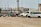 Several vehicles, including vans and SUVs, are parked on a gravel lot. The cars appear to be abandoned or possibly in disrepair, missing wheels, and marked with writing on their windows. In the background, there are industrial buildings and a train on elevated tracks.