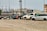 Several vehicles, including vans and SUVs, are parked on a gravel lot. The cars appear to be abandoned or possibly in disrepair, missing wheels, and marked with writing on their windows. In the background, there are industrial buildings and a train on elevated tracks.
