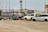 Several vehicles, including vans and SUVs, are parked on a gravel lot. The cars appear to be abandoned or possibly in disrepair, missing wheels, and marked with writing on their windows. In the background, there are industrial buildings and a train on elevated tracks.