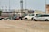Several vehicles, including vans and SUVs, are parked on a gravel lot. The cars appear to be abandoned or possibly in disrepair, missing wheels, and marked with writing on their windows. In the background, there are industrial buildings and a train on elevated tracks.