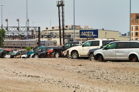 Several vehicles, including vans and SUVs, are parked on a gravel lot. The cars appear to be abandoned or possibly in disrepair, missing wheels, and marked with writing on their windows. In the background, there are industrial buildings and a train on elevated tracks.