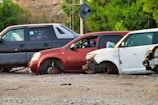 Several cars without wheels and partially stripped of parts are parked on a gravel surface, surrounded by greenery and other discarded items.