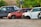 Several cars without wheels and partially stripped of parts are parked on a gravel surface, surrounded by greenery and other discarded items.