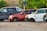 Several cars without wheels and partially stripped of parts are parked on a gravel surface, surrounded by greenery and other discarded items.