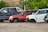 Several cars without wheels and partially stripped of parts are parked on a gravel surface, surrounded by greenery and other discarded items.