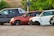 Several cars without wheels and partially stripped of parts are parked on a gravel surface, surrounded by greenery and other discarded items.