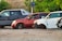 Several cars without wheels and partially stripped of parts are parked on a gravel surface, surrounded by greenery and other discarded items.