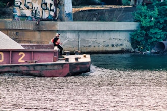 A man wearing a life jacket stands on a barge floating on a river. The barge is carrying a load of gravel and is positioned under a bridge covered with graffiti. There is a concrete wall along the riverbank and dense greenery nearby.