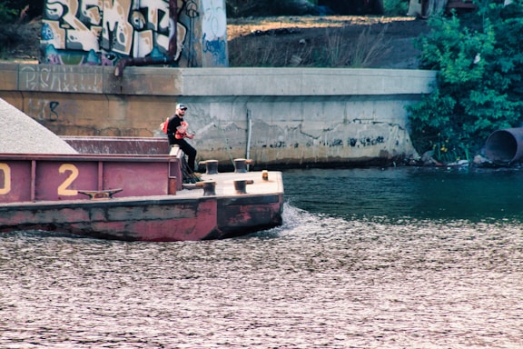 A man wearing a life jacket stands on a barge floating on a river. The barge is carrying a load of gravel and is positioned under a bridge covered with graffiti. There is a concrete wall along the riverbank and dense greenery nearby.