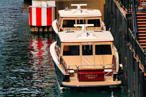 Two medium-sized boats with beige canopies are docked next to a pier. A red and white-striped buoy is visible on the left, and a set of stairs is on the right.