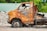 An old, rusted vehicle with missing parts sits abandoned in a scrapyard. The surface of the vehicle is heavily corroded, with peeling paint and rust exposing the metal beneath. Surrounding the vehicle, there are piles of gravel and other scrap materials, while a green corrugated roof is visible in the background through lush foliage.