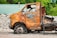 An old, rusted vehicle with missing parts sits abandoned in a scrapyard. The surface of the vehicle is heavily corroded, with peeling paint and rust exposing the metal beneath. Surrounding the vehicle, there are piles of gravel and other scrap materials, while a green corrugated roof is visible in the background through lush foliage.