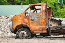An old, rusted vehicle with missing parts sits abandoned in a scrapyard. The surface of the vehicle is heavily corroded, with peeling paint and rust exposing the metal beneath. Surrounding the vehicle, there are piles of gravel and other scrap materials, while a green corrugated roof is visible in the background through lush foliage.