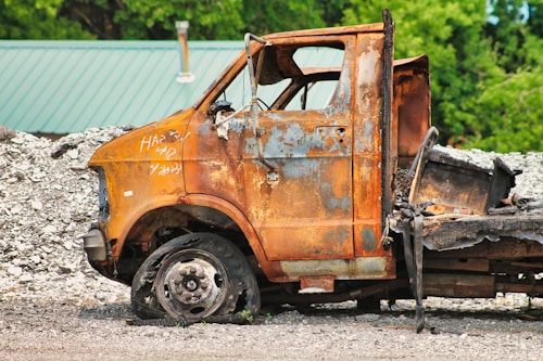 An old, rusted vehicle with missing parts sits abandoned in a scrapyard. The surface of the vehicle is heavily corroded, with peeling paint and rust exposing the metal beneath. Surrounding the vehicle, there are piles of gravel and other scrap materials, while a green corrugated roof is visible in the background through lush foliage.