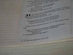 A close-up view of an open book displaying text from a religious or spiritual scripture with numbered verses. The text is formatted with bold numbers indicating specific sections or verses, and includes phrases referencing divine declarations.