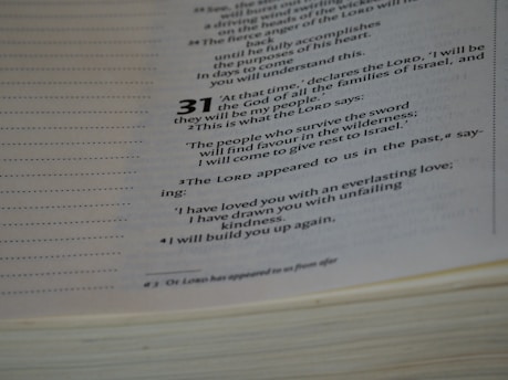 A close-up view of an open book displaying text from a religious or spiritual scripture with numbered verses. The text is formatted with bold numbers indicating specific sections or verses, and includes phrases referencing divine declarations.