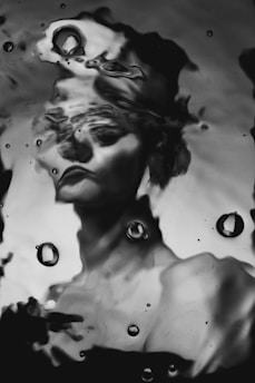 A distorted black and white portrait of a person with ripples and water droplets obscuring the features. The water effect creates an abstract and surreal atmosphere, transforming the facial features into an impressionistic blur.