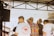 A group of people is gathered in an outdoor setting with a partially covered structure above them. One person wears a colorful patterned shirt, and another person has a shirt with the text 'Bangkok Skate Club' along with an elephant design. There is a fan mounted on the left, and metal beams and railings are visible in the background.