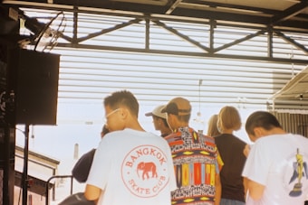 A group of people is gathered in an outdoor setting with a partially covered structure above them. One person wears a colorful patterned shirt, and another person has a shirt with the text 'Bangkok Skate Club' along with an elephant design. There is a fan mounted on the left, and metal beams and railings are visible in the background.