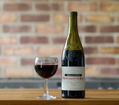 A bottle of Chateauneuf-du-Pape wine is positioned next to a glass filled with red wine, both placed on a wooden surface. The background is a blurred brick wall, offering a rustic and cozy atmosphere.