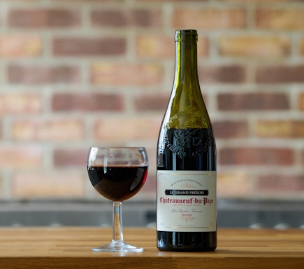 A bottle of Chateauneuf-du-Pape wine is positioned next to a glass filled with red wine, both placed on a wooden surface. The background is a blurred brick wall, offering a rustic and cozy atmosphere.