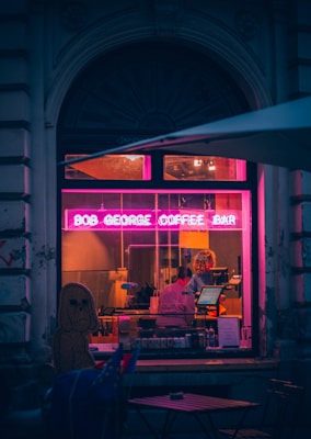 A cozy coffee bar with a vibrant pink neon sign, 'Bob George Coffee Bar', illuminates the front window. Inside, a person stands behind the counter working at a coffee machine. The façade has a rustic, old-world charm with stonework and an arched window. The scene is set in the evening, creating a warm and inviting atmosphere.