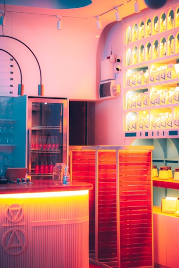 A modern interior with vibrant lighting, featuring shelves with illuminated arches. A counter with decorative patterns and various bottles can be seen. Bright colors create a lively atmosphere.