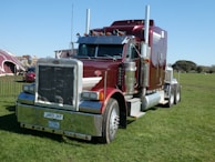 A large, shiny red semi-truck with chrome details is parked on a grassy field. The truck has a prominent front grille, dual exhaust stacks, and large black tires. In the background, there are tents and some trees under a clear blue sky.