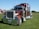 A large, shiny red semi-truck with chrome details is parked on a grassy field. The truck has a prominent front grille, dual exhaust stacks, and large black tires. In the background, there are tents and some trees under a clear blue sky.