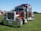 A large, shiny red semi-truck with chrome details is parked on a grassy field. The truck has a prominent front grille, dual exhaust stacks, and large black tires. In the background, there are tents and some trees under a clear blue sky.