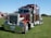 A large, shiny red semi-truck with chrome details is parked on a grassy field. The truck has a prominent front grille, dual exhaust stacks, and large black tires. In the background, there are tents and some trees under a clear blue sky.