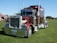 A large, shiny red semi-truck with chrome details is parked on a grassy field. The truck has a prominent front grille, dual exhaust stacks, and large black tires. In the background, there are tents and some trees under a clear blue sky.