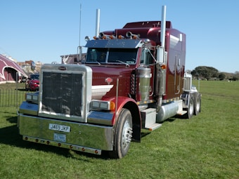 A large, shiny red semi-truck with chrome details is parked on a grassy field. The truck has a prominent front grille, dual exhaust stacks, and large black tires. In the background, there are tents and some trees under a clear blue sky.