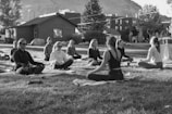 A group of people are sitting cross-legged on yoga mats in an outdoor setting, likely a park or open area. They appear to be engaged in a meditation or yoga session, with a serene and calm demeanor. The setting includes a few trees, a wooden building in the background, and a distant view of a hillside. The image is in black and white, adding a timeless and tranquil feel to the scene.