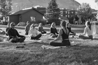 A group of people are sitting cross-legged on yoga mats in an outdoor setting, likely a park or open area. They appear to be engaged in a meditation or yoga session, with a serene and calm demeanor. The setting includes a few trees, a wooden building in the background, and a distant view of a hillside. The image is in black and white, adding a timeless and tranquil feel to the scene.