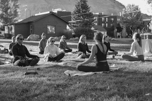 A group of people are sitting cross-legged on yoga mats in an outdoor setting, likely a park or open area. They appear to be engaged in a meditation or yoga session, with a serene and calm demeanor. The setting includes a few trees, a wooden building in the background, and a distant view of a hillside. The image is in black and white, adding a timeless and tranquil feel to the scene.