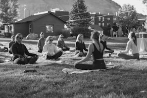 A group of people are sitting cross-legged on yoga mats in an outdoor setting, likely a park or open area. They appear to be engaged in a meditation or yoga session, with a serene and calm demeanor. The setting includes a few trees, a wooden building in the background, and a distant view of a hillside. The image is in black and white, adding a timeless and tranquil feel to the scene.
