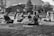 A group of people are sitting cross-legged on yoga mats in an outdoor setting, likely a park or open area. They appear to be engaged in a meditation or yoga session, with a serene and calm demeanor. The setting includes a few trees, a wooden building in the background, and a distant view of a hillside. The image is in black and white, adding a timeless and tranquil feel to the scene.