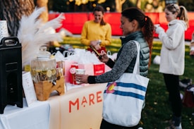 A group of people is gathered around a table set up outdoors, featuring a beverage dispenser with lemon water, paper cups, and various gift bags. One woman in a green jacket and white tote bag with a colorful design is holding a cup and interacting with her phone. Another person in a yellow top and another in a white jacket are also visible in the background. The setting appears to be a park with green grass and a red backdrop.