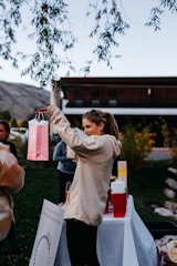 A person in a light hoodie holds up a colorful gift bag. Tables are set up outdoors with more gift bags and informational signs. A few people are gathered in the background, surrounded by early autumn foliage and a building.