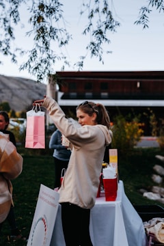 A person in a light hoodie holds up a colorful gift bag. Tables are set up outdoors with more gift bags and informational signs. A few people are gathered in the background, surrounded by early autumn foliage and a building.
